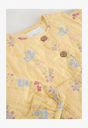Kollane tikitud jakk, millel on lillemuster siniste ja roosade lilledega, puidust nööbid ning tekstureeritud kangas õmblusega detailidega.