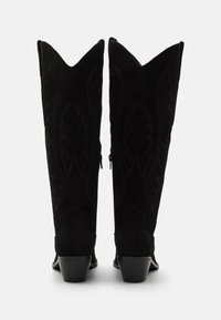 Botas de vaquero de ante negro con puntas afiladas, costuras decorativas, cremalleras laterales y tacones apilados. Acabado texturizado con un diseño elegante.