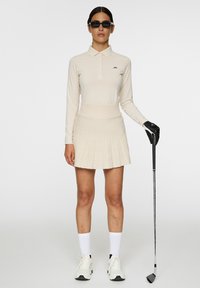 Beige Langarm-Polo, plissierter beiger Rock, schwarze Sonnenbrille, weiße Socken und weiße Sportschuhe. Hält einen Golfschläger in einer Hand.