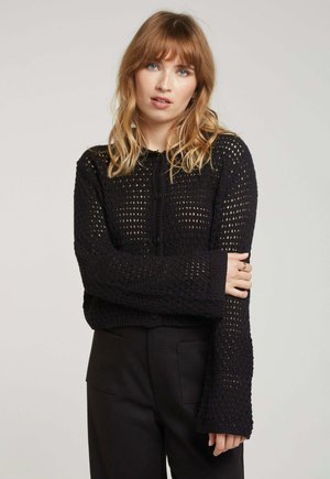Cardigan noir en tricot ouvert avec de longues manches et un décolleté rond, présentant des fermetures à boutons et un motif texturé. Porté par-dessus un pantalon noir.