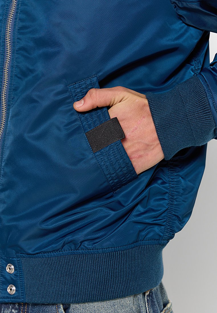 Veste bomber bleu marine avec une texture lisse et brillante, des poignets côtelés et une poche latérale avec un rabat en velcro noir.