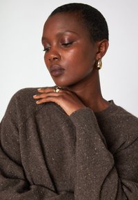 Gros plan sur une femme aux cheveux courts portant un pull marron à taches, des boucles d'oreilles en or et une bague en or, posant les yeux fermés.