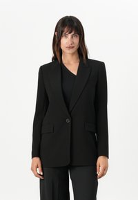 Blazer nera su misura con rever a punta, chiusura con un bottone, due tasche frontali e una texture di tessuto liscia.