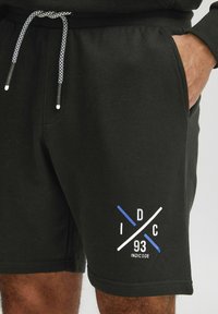 Sorte bomulds shorts med elastisk talje, prydet med et hvidt og blåt grafisk design nederst til venstre, samt snoretræk med sort og hvide detaljer.