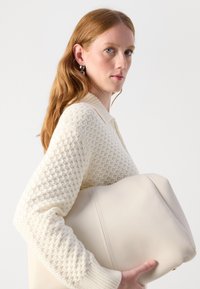 Borsa a mano in pelle color crema con un design minimalista, tenuta contro un maglione in tessuto cashmere avorio con colletto a costine.