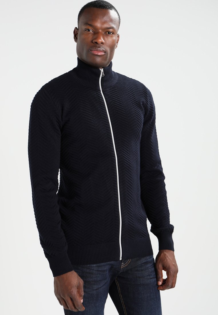 Marineblåe zip-jakke med teksturert sikksakkmønster, høy krage og kontrastfarget hvit glidelås. Ribbestrikkede mansjetter og kant; tettsittende design.