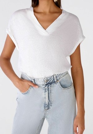 Femme portant un haut blanc à manches courtes en V, rentré dans un jean taille haute bleu clair, une main dans la poche.