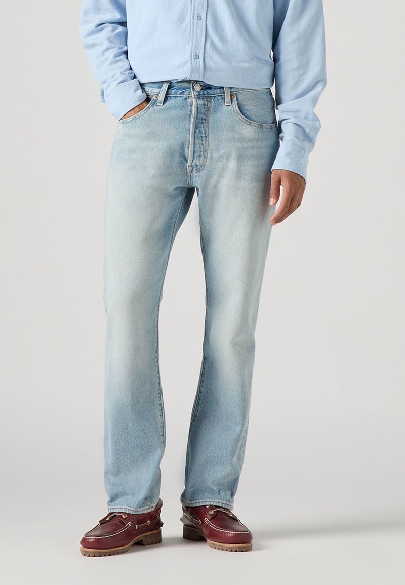 Homme portant un jean bleu clair, une chemise bleu clair à manches longues et des chaussures en cuir marron, avec une main dans la poche et l'autre détendue.