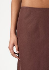 Brown, textured fabric skirt, close-up view of the hem and hand resting on it, showcasing simple design and clean stitching.