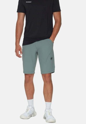 Hombre vistiendo camiseta negra de Mammut, pantalones cortos azules claros hasta la rodilla con logo en la pierna izquierda, calcetas blancas y zapatos deportivos blancos, de pie con una mano en el bolsillo.