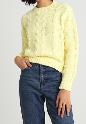 Femme portant un pull en maille torsadée jaune pâle et un jean bleu foncé taille haute, une main dans la poche et l'autre reposant le long du corps.