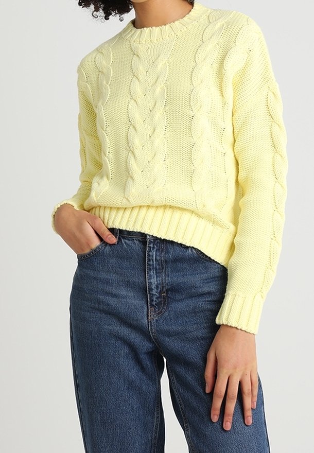 Femme portant un pull en maille torsadée jaune pâle et un jean bleu foncé taille haute, une main dans la poche et l'autre reposant le long du corps.