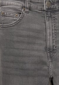 Gros plan d'un jean en denim gris montrant la poche avant, le bouton métallique, la passants de ceinture et les détails de couture.