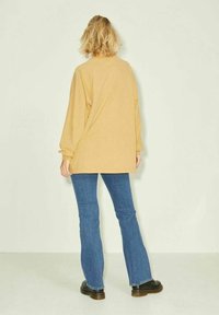 Maglietta gialla in cotone a maniche lunghe e oversize con collo rotondo, abbinata a jeans svasati blu chiaro e calzature marroni scure.