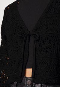Černý pletený cardigan s otevřeným designem, zdobený texturovaným vzorem, s zavazováním vpředu a rukávy s délkou tří čtvrtin.