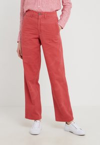 Osoba nosící červené kalhoty s vysokým pasem a širokými nohavicemi se bočními kapsami, v kombinaci s bílými teniskami a dlouhým rukávem růžového pruhovaného trička.