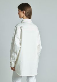 Camisa blanca de manga larga con cuello puntiagudo y bordado floral en los hombros, con un corte holgado y un dobladillo curvado.