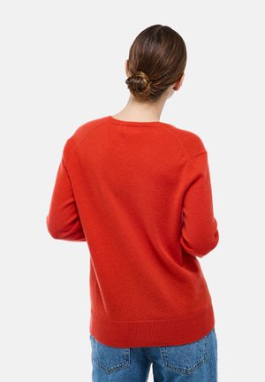 Personne avec des cheveux bruns en chignon portant un pull rouge vif et un jean bleu, vue de dos sur un fond uni.