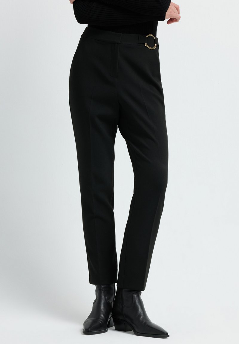 Vrouw in zwarte taps toelopende broek met een gouden gespdetail en zwarte puntige enkellaarsjes, staand tegen een effen lichte achtergrond.