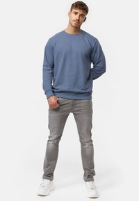Sudadera azul clara con puños y dobladillo acanalados, cuello redondo, combinada con vaqueros grises y zapatillas blancas. Sin logotipos ni patrones visibles.