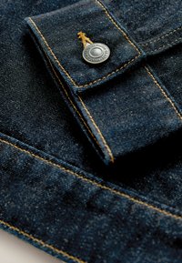 Primer plano de tela vaquera de color azul oscuro, con costuras amarillas contrastantes y un botón metálico plateado con texto en relieve.