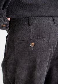Black corduroy pants with ribbed texture, featuring a back pocket with a brown button closure and a tailored fit.