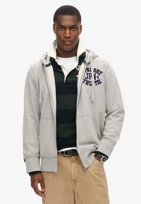Uomo che indossa pantaloni beige, cintura nera, polo a righe verdi e nere sotto una felpa grigia chiara con zip aperta e scritta sul petto, mano nella tasca.
