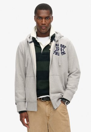 Hombre con pantalones beige, cinturón negro, camiseta polo a rayas verdes y negras debajo de una sudadera con cremallera gris claro abierta, con texto en el pecho, con una mano en el bolsillo.