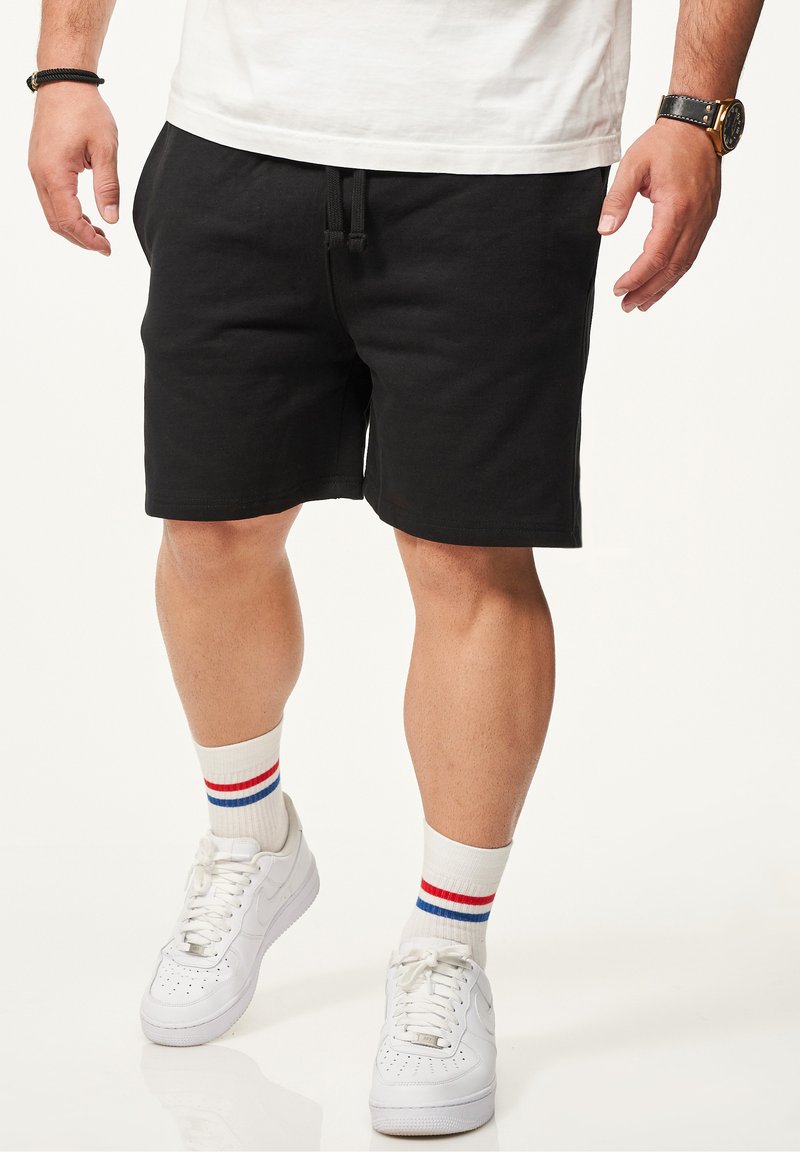 Schwarze Baumwollshorts mit elastischem Bund und Kordelzug, kombiniert mit weißen Sneakers und gestreiften weißen Socken mit roten und blauen Akzenten.