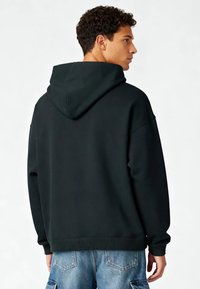 Sweat à capuche noir avec une grande capuche, épaules tombantes et poignets côtelés. Texture douce avec une coupe ample, porté avec un short en jean bleu clair.
