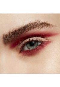 Gros plan d'un œil avec une paupière beige clair et un fard à paupières rouge vif s'étendant au-delà du pli, encadré par des cils épais et foncés ainsi que des sourcils.