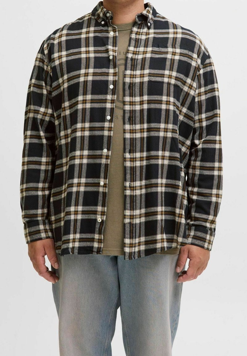 Homme portant une chemise à carreaux noire, blanche et marron déboutonnée sur un t-shirt graphique vert avec un jean bleu clair sur un fond neutre.