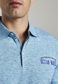 Světle modrá polokošile se strukturovanou tkaninou, krátkým límcem a třemi bílými knoflíky. Má vyšívaný nápis "OCEAN WAVES" tmavě modrou barvou.