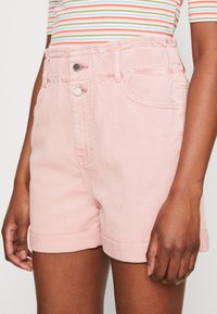 Vaaleanpunaiset farkkushortsit, joissa on kaksi etupussia, käännetyt lahkeet ja hopeiset yksityiskohdat vyötäröllä, esittelevät pehmeää kangastuntumaa.