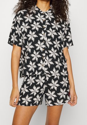 Mujer vestida con una camisa negra de manga corta abotonada y pantalones cortos a juego con un patrón de flores blancas.