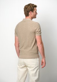 Beige short-sleeve polo shirt in textured knit, featuring a classic collar and ribbed hem, worn with light-colored trousers.