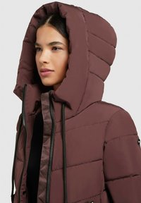 Bordová péřová bunda s velkou kapucí, s vodorovnými prošívanými sekcemi, černým zipem a detaily na stahovací šňůrce. Měkká textura.