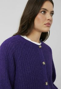 Cardigan violet en tricot avec un motif texturé, doté de boutons métalliques et d'un col arrondi. Le tissu semble épais et confortable.