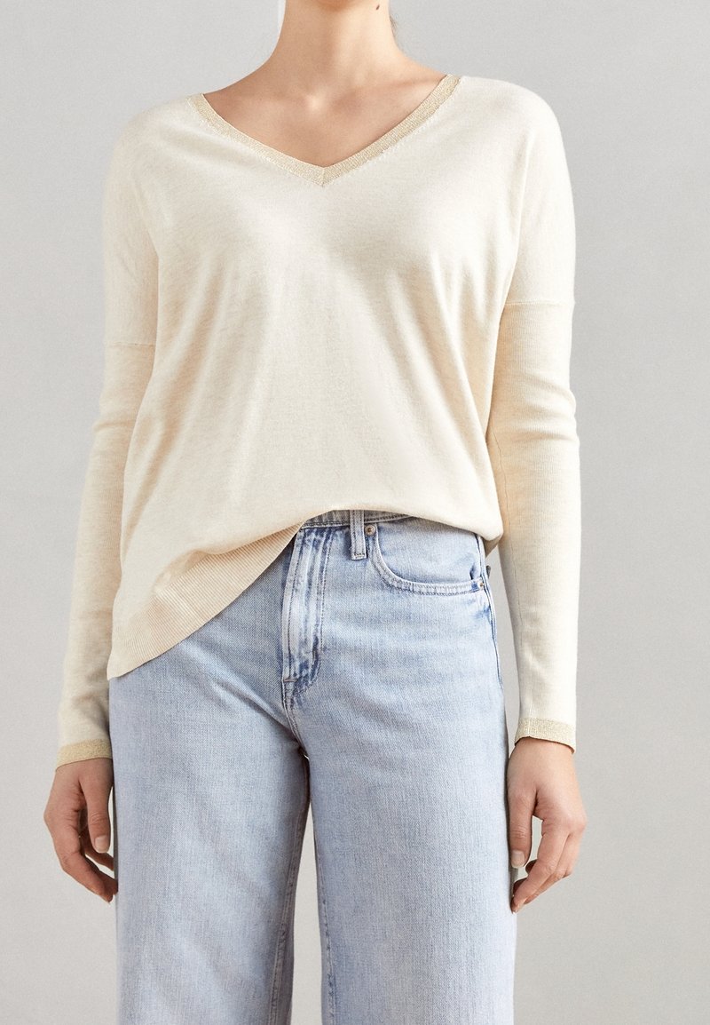 Personne portant un pull crème à col en V manches longues rentré dans un jean bleu clair taille haute sur un fond uni.