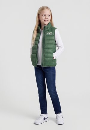 Ragazza che indossa un gilet imbottito verde, una maglia a maniche lunghe bianca, jeans blu e scarpe da ginnastica bianche, in piedi con le mani in tasca e sorridente.