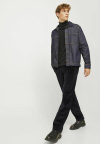 Denim jakna tamno plave boje s kontrastnim narančastim šavovima, preko crne dolčevite, u kombinaciji s crnim kordura hlačama i crnim patikama.