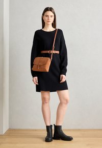 Donna che indossa un vestito maglione nero con cintura marrone, una borsa a tracolla marrone abbinata e stivali neri alla caviglia, in piedi su un pavimento di legno.