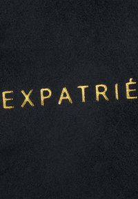 Fekete velúr anyag, arany színű hímzéssel az "EXPATRIÉ" szóval, sima textúrával és egyszerű, modern tipográfiával.