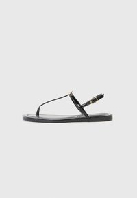 Sandale din piele patent neagră cu design cu brâu în T, curea ajustabilă la gleznă și talpă plată. Caracteristici un accent cu cataramă aurie.