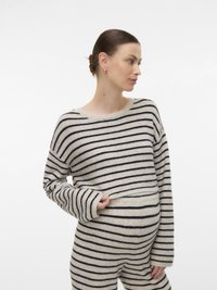 Γυναίκα με μαλλιά σε κότσο, φοράει μπεζ και μαύρο ριγέ πουλόβερ και παντελόνι, σηκώνει απαλά το πουλόβερ για να αποκαλύψει την εγκυμονούσα κοιλιά της, σε λευκό φόντο.