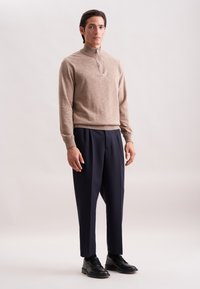 Mann steht in einem beigefarbenen Halbreißverschluss-Pullover, marineblauen, faltigen Hosen und schwarzen Leder-Schuhen auf neutralem Hintergrund.