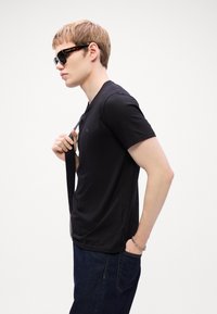 Black short-sleeved t-shirt, smooth texture, paired with dark denim jeans. Model wears sunglasses, holding a black strap over his shoulder.