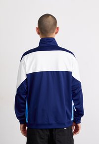 Jaqueta desportiva azul-marinho com painéis brancos e azul-claro, gola alta, mangas longas e textura suave. Apresenta um logotipo perto da bainha.