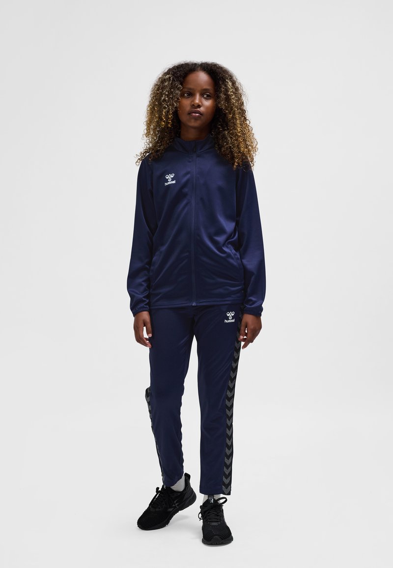 Navy tracksuit with zippered hoodie and pants, featuring a logo on the chest and chevron stripes on the sides. Black athletic shoes.