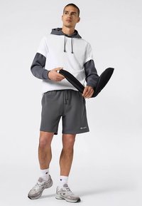 Sudadera con capucha a bloques de color gris y blanco con cordones, pantalones cortos grises con logo y zapatillas grises. Modelo sosteniendo dos objetos negros.